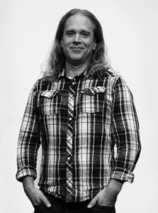 svart vit bild på man med långt hår som står med händerna i fickorna. Rutig skjorta.